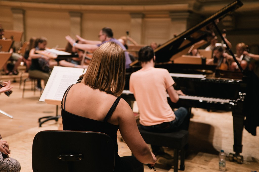 A view of a music rehearsal featuring a pianist and musicians in an orchestra setting, with people playing various instruments and sheet music on stands.